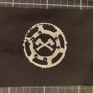 May include: A black fabric patch with a white circular design. The design features two crossed axes inside a circle with a textured border.