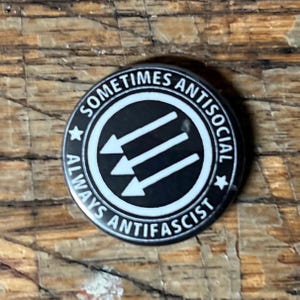Op de afbeelding: Een ronde, zwart-witte knoop met de tekst "Sometimes Antisocial Always Antifascist" rondom een centraal ontwerp van drie witte pijlen naar beneden. De knoop ligt op een houten achtergrond.