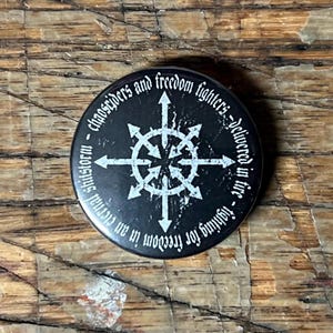 May include: A black button with white text and a white symbol in the center. The text reads "chaosriders and freedom fighters - delivered in fire - fighting for freedom in an eternal shitstorm."