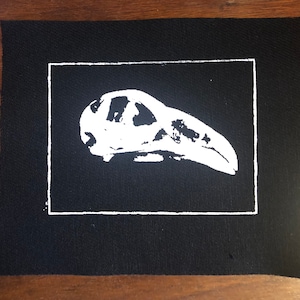 May include: A black fabric patch with a white border and a white image of a bird skull inside.