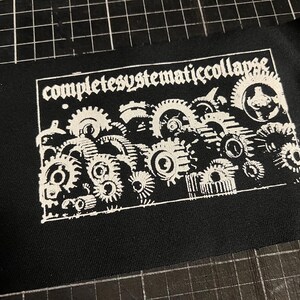 以下が含まれることがあります： 黒い生地のパッチ。ゴシック体の「completesystematiccollapse」の文字が書かれています。白い歯車が組み合わさったデザインで、機械的なテーマを表現しています。パッチは長方形です。