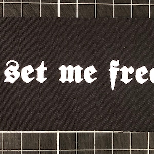 May include: Black fabric patch with white text in a gothic font that reads "Set me free".