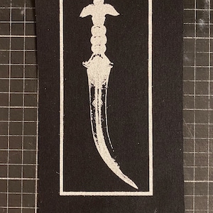 以下が含まれることがあります： 黒い布のパッチに白い刺繍の短剣が施されています