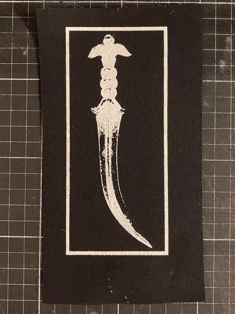 K&ouml;nnte beinhalten: Wei&szlig; gestickter Dolch auf einem schwarzen Stoff-Patch.