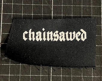 May include: A black fabric patch with the word "chainsawed" in white gothic lettering. The patch is rectangular with slightly frayed edges, set against a black grid background.