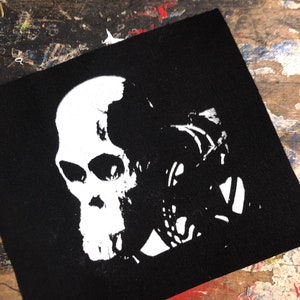 Puede incluir: Parche cuadrado negro con un diseño de calavera blanca. La calavera tiene cuencas de los ojos detalladas y una mandíbula prominente. El parche es probablemente de tela y adecuado para coser o planchar en ropa o accesorios.
