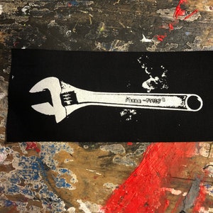 Könnte beinhalten: Weißer Schraubenschlüssel, der auf einem schwarzen Stoff-Patch gedruckt ist. Der Schraubenschlüssel trägt den Aufdruck "Made in USA".