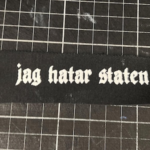 Könnte beinhalten: Schwarzer Stoff-Patch mit weißem Text, der "jag hatar staten" lautet.