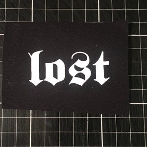 Może przedstawiać: Czarny materiałowy znaczek z napisem "lost" w białym kolorze w stylu gotyckim.
