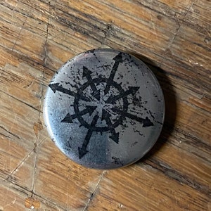 Op de afbeelding: Een zilveren metalen knoop met een zwart-witte afbeelding van een symbool met pijlen die naar buiten wijzen.