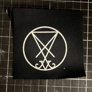 Könnte beinhalten: Ein quadratischer, schwarzer Aufnäher mit einem weißen Lucifer-Sigill, einem Symbol aus der Okkultismus. Das Sigill ist ein umgekehrtes Pentagramm in einem Kreis, mit zusätzlichen Symbolen. Der Aufnäher ist für Kleidung oder Accessoires bestimmt.