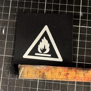 K&ouml;nnte beinhalten: Ein schwarzer quadratischer Aufn&auml;her mit einem wei&szlig;en Dreieck, das ein Flammensymbol enth&auml;lt. Der Aufn&auml;her ist etwa 6,35 cm breit. Das Bild ist auf einer schwarzen Rasteroberfl&auml;che aufgenommen.