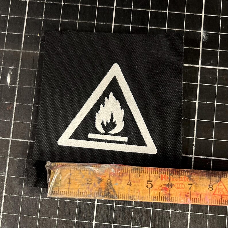 K&ouml;nnte beinhalten: Ein schwarzer quadratischer Aufn&auml;her mit einem wei&szlig;en Dreieck, das ein Flammensymbol enth&auml;lt. Der Aufn&auml;her ist etwa 6,35 cm breit. Das Bild ist auf einer schwarzen Rasteroberfl&auml;che aufgenommen.