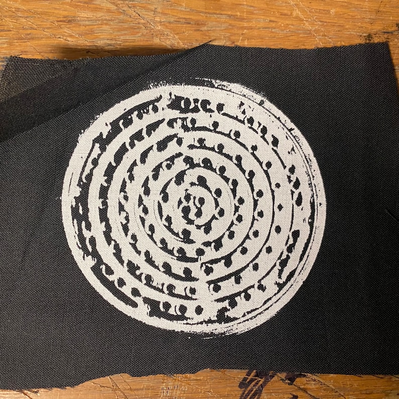 Pode incluir: Um design circular branco sobre um fundo de tecido preto. O design &eacute; composto por c&iacute;rculos conc&ecirc;ntricos de pequenos pontos.