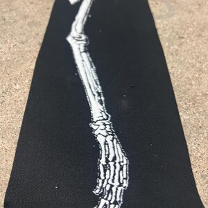K&ouml;nnte beinhalten: Ein schwarzer, rechteckiger Aufn&auml;her mit einer wei&szlig;en Illustration eines Skelettarms und einer Hand. Die detaillierte Abbildung zeigt die Knochen von Arm und Hand. Der Aufn&auml;her ist aus einem dunklen Stoff gefertigt.