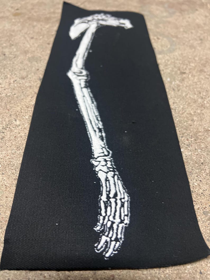 K&ouml;nnte beinhalten: Ein schwarzer, rechteckiger Aufn&auml;her mit einer wei&szlig;en Illustration eines Skelettarms und einer Hand. Die detaillierte Abbildung zeigt die Knochen von Arm und Hand. Der Aufn&auml;her ist aus einem dunklen Stoff gefertigt.