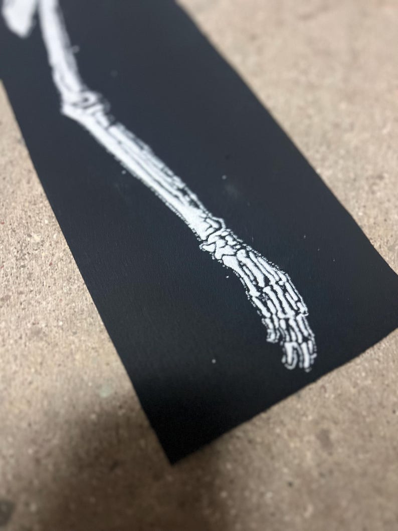 K&ouml;nnte beinhalten: Eine schwarze rechteckige Karte zeigt eine wei&szlig;e Illustration eines menschlichen Arm- und Handskeletts. Das Skelettdesign ist detailliert, mit sichtbaren Knochen und Gelenken. Das Bild ist eine Nahaufnahme, die den Kontrast zwischen der wei&szlig;en Illustration und dem schwarzen Hintergrund hervorhebt.