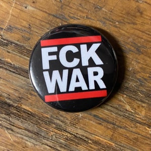Może przedstawiać: Czarny guzik z czerwonymi i białymi paskami. Guzik ma napis "FCK WAR" białymi literami.