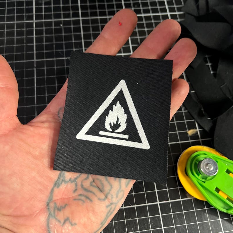 K&ouml;nnte beinhalten: Ein schwarzer quadratischer Aufn&auml;her mit einem wei&szlig;en Dreieck, das ein Flammensymbol enth&auml;lt. Der Aufn&auml;her wird in einer Hand gehalten, mit einem gr&uuml;n-gelben Rollschneider im Hintergrund. Der Aufn&auml;her ist wahrscheinlich ein Warnhinweis.