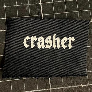 Op de afbeelding: Een zwart stoffen label met het woord "crasher" gedrukt in witte gotische letters. Het label heeft een getextureerd oppervlak en een rechthoekige vorm. De achtergrond is een zwarte snijmat met een rasterpatroon.