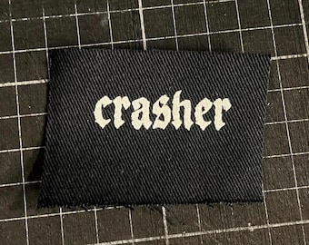 May include: A black fabric label with the word "crasher" printed in white gothic lettering. The label has a textured surface and a rectangular shape. The background is a black cutting mat with a grid pattern.