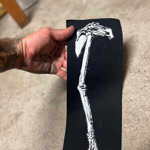 K&ouml;nnte beinhalten: Schwarzer rechteckiger Aufn&auml;her mit einem wei&szlig;en Skelettarm- und Schulterblatt-Design. Der Aufn&auml;her wird vor einer Betonoberfl&auml;che gehalten. Das Design ist eine detaillierte Illustration des menschlichen Skeletts.