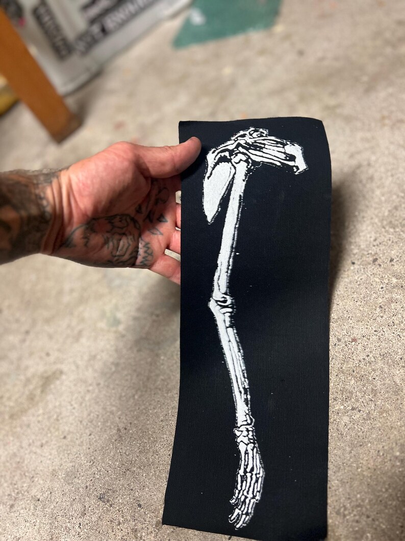 K&ouml;nnte beinhalten: Schwarzer rechteckiger Aufn&auml;her mit einem wei&szlig;en Skelettarm- und Schulterblatt-Design. Der Aufn&auml;her wird vor einer Betonoberfl&auml;che gehalten. Das Design ist eine detaillierte Illustration des menschlichen Skeletts.