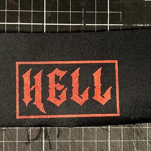 Könnte beinhalten: Schwarzer rechteckiger Aufnäher mit dem Wort "HELL" in roter gotischer Schrift. Der Text ist von einem roten Rahmen umgeben. Der Aufnäher ist für Kleidung oder Accessoires bestimmt.