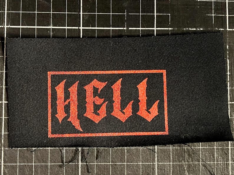May include: Black rectangular patch with the word "HELL" in red gothic lettering. The text is enclosed within a red rectangular border. The patch is likely for clothing or accessories.