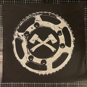 May include: A black fabric patch with a white circular design. The design features two crossed axes inside a gear-like shape.