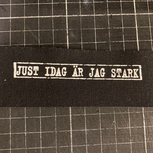May include: Black fabric patch with white text that reads "JUST IDAG ÄR JAG STARK" in a rectangular frame.