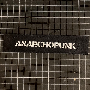 Peut inclure: Un patch en tissu noir avec le texte blanc "ANARCHOPUNK" imprimé dessus.