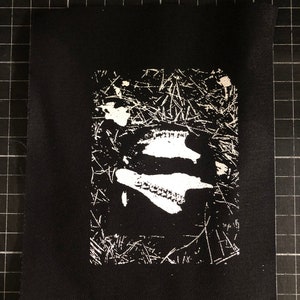 May include: Black fabric patch featuring a white graphic print. The graphic depicts a shoe or boot in a grassy setting. The patch is rectangular and has a minimalist design, suitable for clothing or accessories.