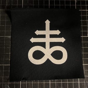 May include: A black fabric patch with a white Leviathan cross emblem. The symbol is a double cross with arrows and an infinity symbol. The patch is square and designed to be attached to clothing or accessories, possibly by sewing or ironing.