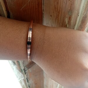 May include: A copper bracelet with black accents. The bracelet is thin and has a simple design.
