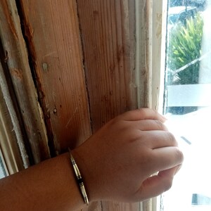 May include: A silver bracelet with a thin, flat design and two black lines running down the center. The bracelet is worn on a person's wrist.
