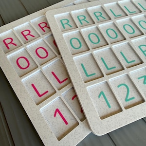 May include: Two rectangular off-white speckled boards featuring a grid of recessed squares. Each square contains a raised letter or number. One board displays pink characters including R, O, L, and 1, while the other features teal characters including R, L, O, 1, and 2.