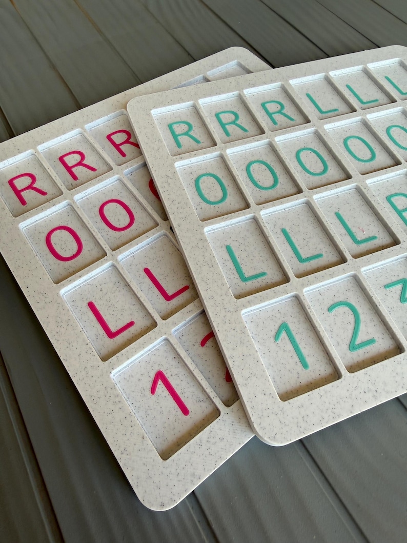 May include: Two rectangular off-white speckled boards featuring a grid of recessed squares. Each square contains a raised letter or number. One board displays pink characters including R, O, L, and 1, while the other features teal characters including R, L, O, 1, and 2.