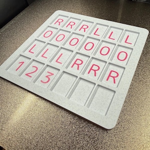 May include: A square, light gray speckled board featuring a grid of recessed rectangular slots. The slots contain pink printed characters arranged in rows: R, R, R, L, L, L in the first row; O, O, O, O, O, O in the second; L, L, L, R, R, R in the third; and 1, 2, 3 in the fourth.