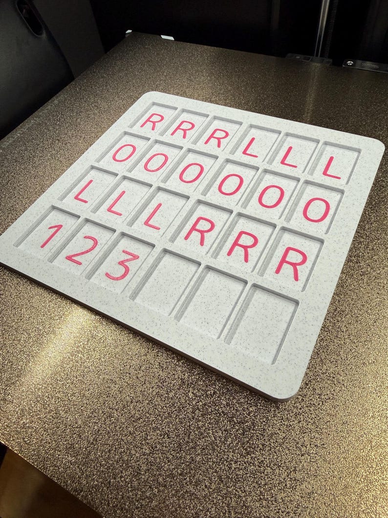 May include: A square, light gray speckled board featuring a grid of recessed rectangular slots. The slots contain pink printed characters arranged in rows: R, R, R, L, L, L in the first row; O, O, O, O, O, O in the second; L, L, L, R, R, R in the third; and 1, 2, 3 in the fourth.
