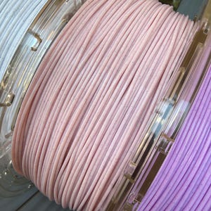 May include: Three plastic spools of 3D printer filament wound with thin, solid-colored strands. The center spool is light pink, the left is white, and the right is light purple. Each strand features a subtle, speckled glitter texture throughout.