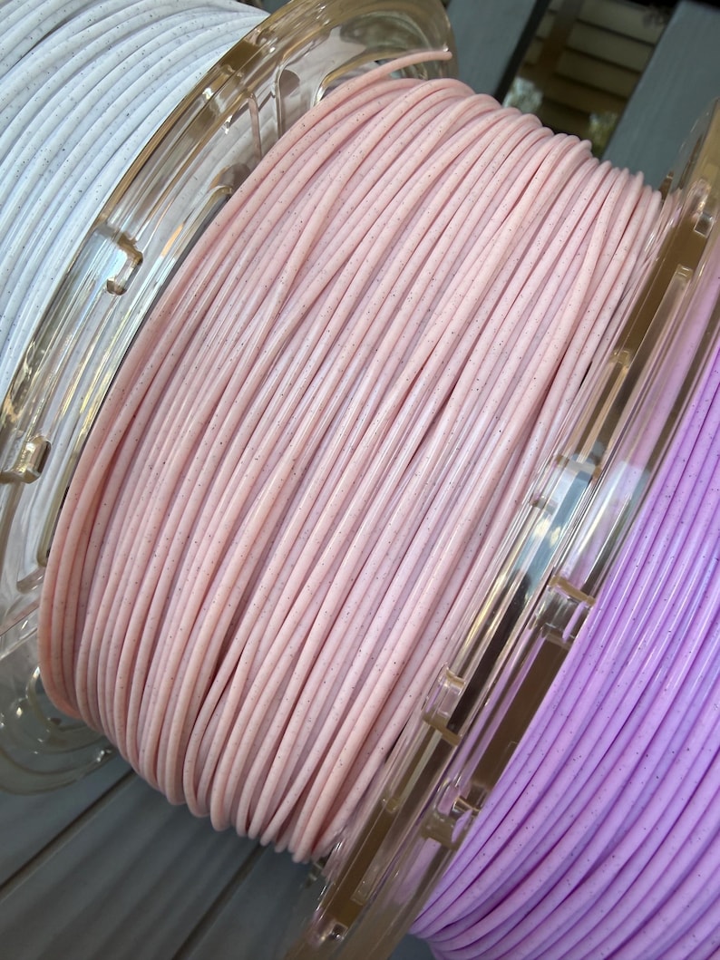 May include: Three plastic spools of 3D printer filament wound with thin, solid-colored strands. The center spool is light pink, the left is white, and the right is light purple. Each strand features a subtle, speckled glitter texture throughout.
