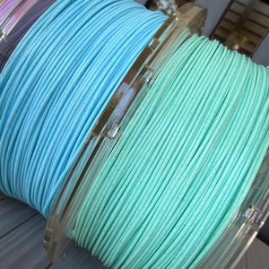 May include: Three circular spools of 3D printer filament arranged in a row. The spools feature light purple, bright sky blue, and soft mint green filament, each with a subtle speckled texture throughout the wound plastic material.