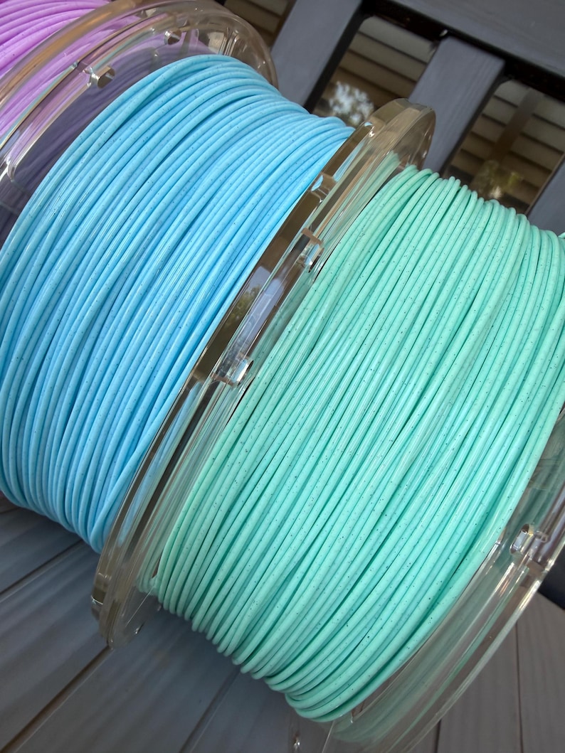 May include: Three circular spools of 3D printer filament arranged in a row. The spools feature light purple, bright sky blue, and soft mint green filament, each with a subtle speckled texture throughout the wound plastic material.