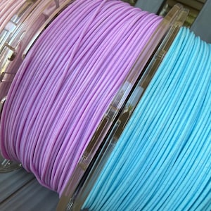 May include: Three spools of 3D printer filament in pastel shades of pink, lavender, and light blue. Each spool features a clear plastic core and contains a continuous strand of filament with a subtle speckled texture throughout the material.