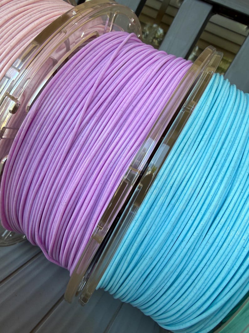 May include: Three spools of 3D printer filament in pastel shades of pink, lavender, and light blue. Each spool features a clear plastic core and contains a continuous strand of filament with a subtle speckled texture throughout the material.
