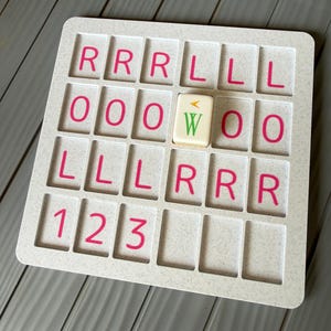 May include: A white speckled plastic grid board with four rows of rectangular slots. The slots contain pink printed letters R, L, O and numbers 1, 2, 3. A single cream-colored tile with a green letter W and a small orange arrow sits in one slot.