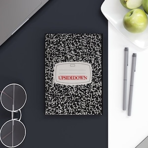 Puede incluir: Un cuaderno de composición negro y blanco con la palabra "UPSIDEDOWN" impresa en rojo en la portada. El cuaderno está sobre una superficie negra junto a un ordenador portátil y un par de gafas redondas.