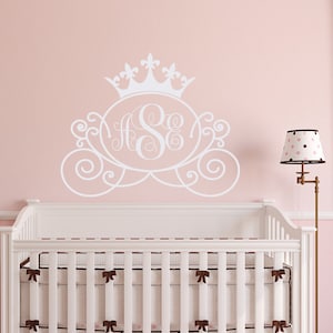 May include: White crib with a pink polka dot skirt and a white crown and carriage monogram decal on the wall behind it.