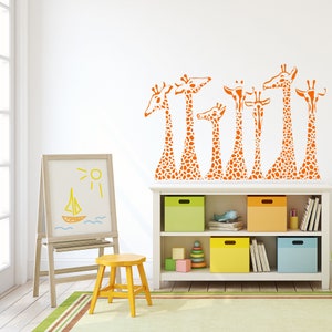 May include: A white wall with a decal of orange giraffes. A white bookshelf with colorful storage bins and toys sits below the decal. A white easel with a drawing of a sailboat and sun sits to the left of the bookshelf.